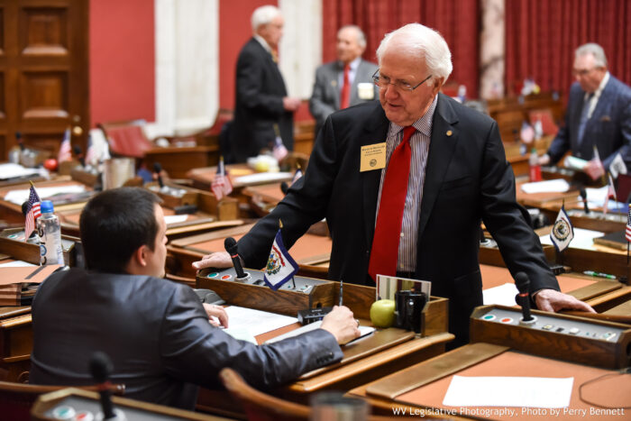 Man with white Hair, red tie, blue suit talking to another man sitting down.