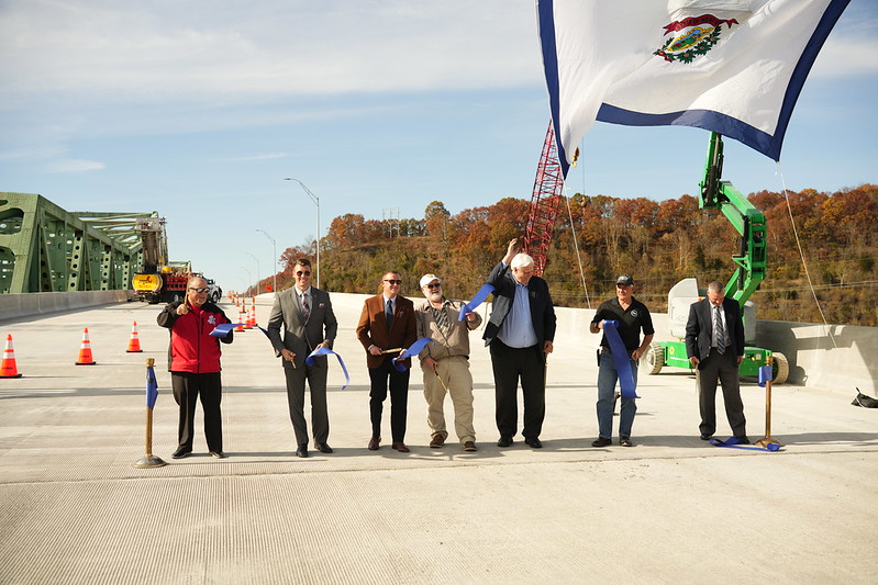 A group of men stands in a line on a concrete bridge, cutting a blue ribbon on a crisp fall day with the state flag flying above them.