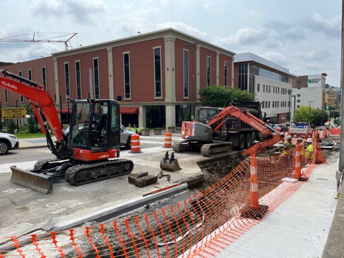 Construction machinery crowds a busy street in a city's central business district with orange cones blocking off a trench between the street and the sidewalk.