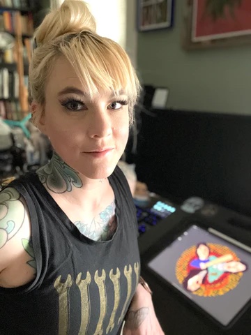 Woman takes a selfie. Artwork is behind her on a table. She has blonde hair that is pulled up into a messy bun. She has tattoos on her neck.