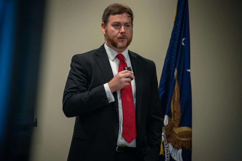 A man in a red tie and formal suit holds a microphone. A flag is behind him.