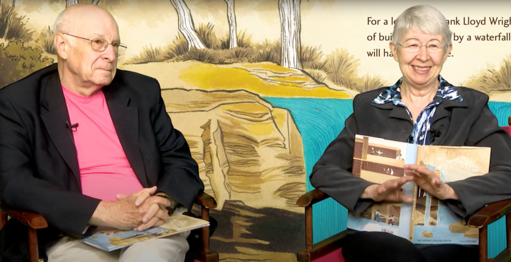 Two older people -- a woman and a man -- sit next to each other for a video interview. The man is wearing a pink shirt and black jacket, while the woman wears a black jacket and blue shirt. Both wear glasses. The woman holds a children's book with the title "Fallingwater."