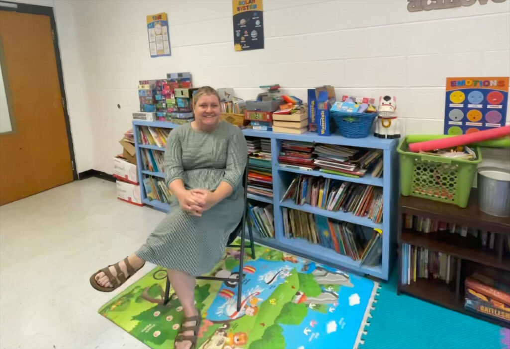 Woman sits in children's classroom wearing long dress and sandals.