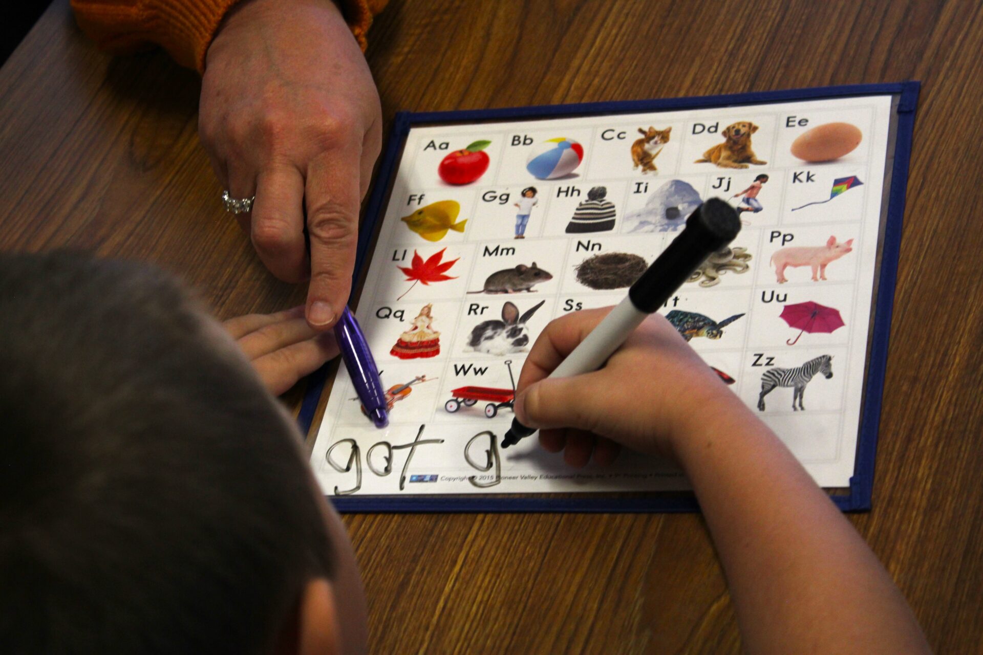 An adult hand indicates a letter written by a student on a letter/image board. The child can be seen holding a dry erase marker and writing the word "got" for a second time at the bottom of the board, where short words are paired with simple images. The entire vignette is seen over the child's shoulder.