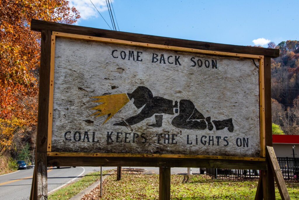 A handmade sign is shown of a coal miner crawling through a crawl space with a helmet on. The sign reads, "Come back soon. Coal keeps the lights on."