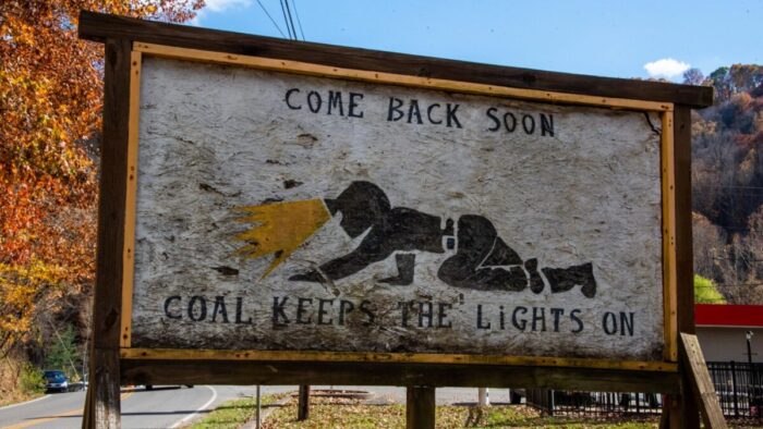A handmade sign is shown of a coal miner crawling through a crawl space with a helmet on. The sign reads, "Come back soon. Coal keeps the lights on."