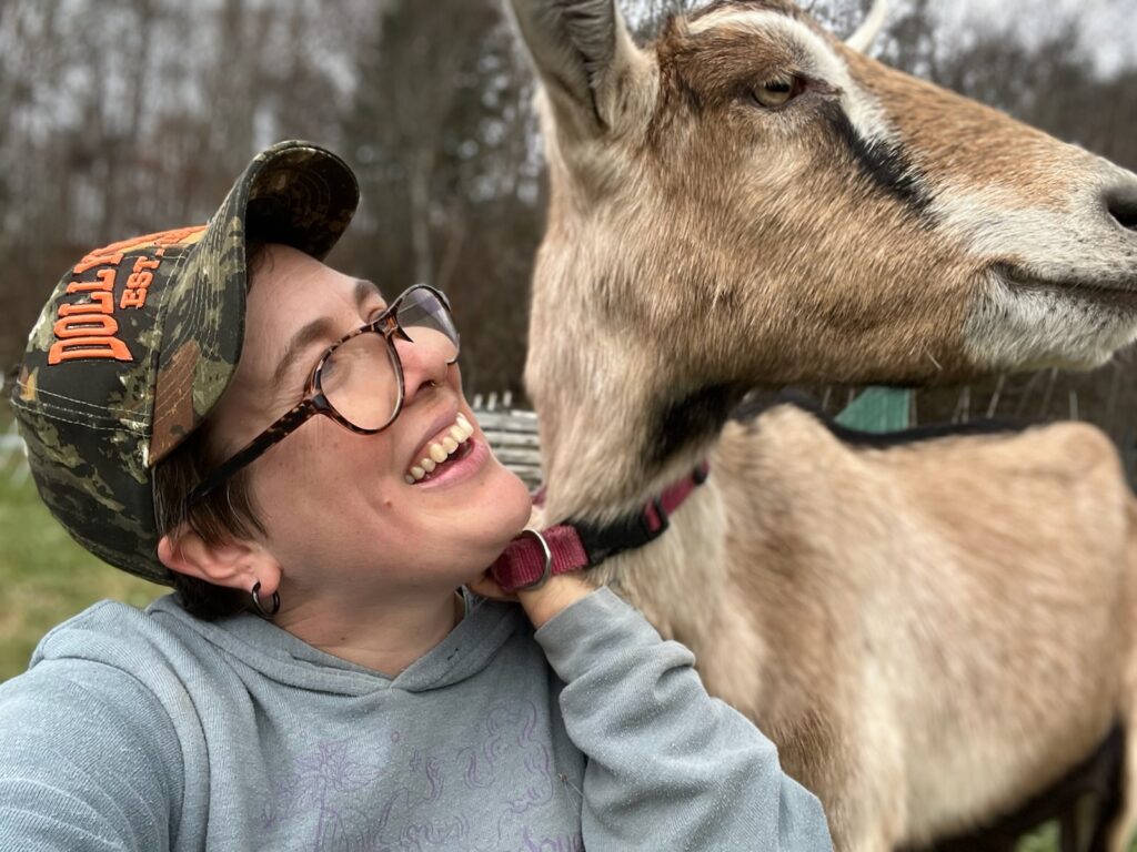 A person wearing a ball cap, glasses, and a gray sweatshirt poses with their pet goat. The person is smiling up at the goat. The goat has brown fur.