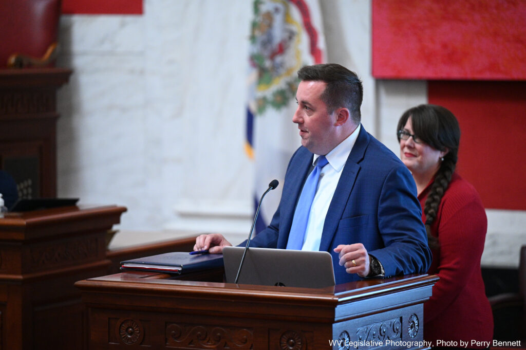 A man in a dark blue suit, white shirt, and powder blue tie speaks at a wooden lectern in the West Virginia Senate Chamber. Behind him can be seen a marble wall with a West Virginia state flag, as well as a woman wearing glasses and a red sweater dress.