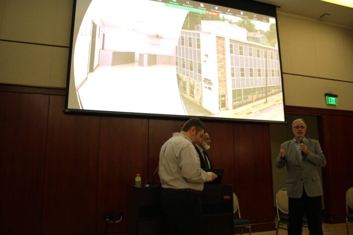 Three men stand under a screen in a paneled room . The man on the right wears a grey jacket while speaking into a microphone. The screen displays the interior and exterior of a building.