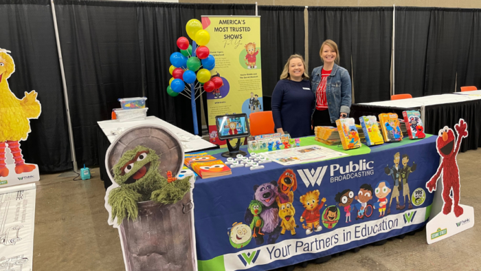 Two women stand next to each other behind a table smiling for the camera. There are cut out standees of Sesame Street characters around the table as well as colorful balloons.