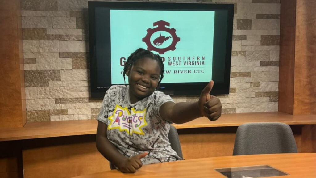 Naveah Woods, a Mercer County student attends the first GEAR UP SWV Media Camp hosted at Concord University in July 2023. She sits at a light wooden desk in a studion while giving a thumbs up to the camera. Behind her can be seen the GEAR UP SWV logo on a screen mounted to a grey tiled wall.