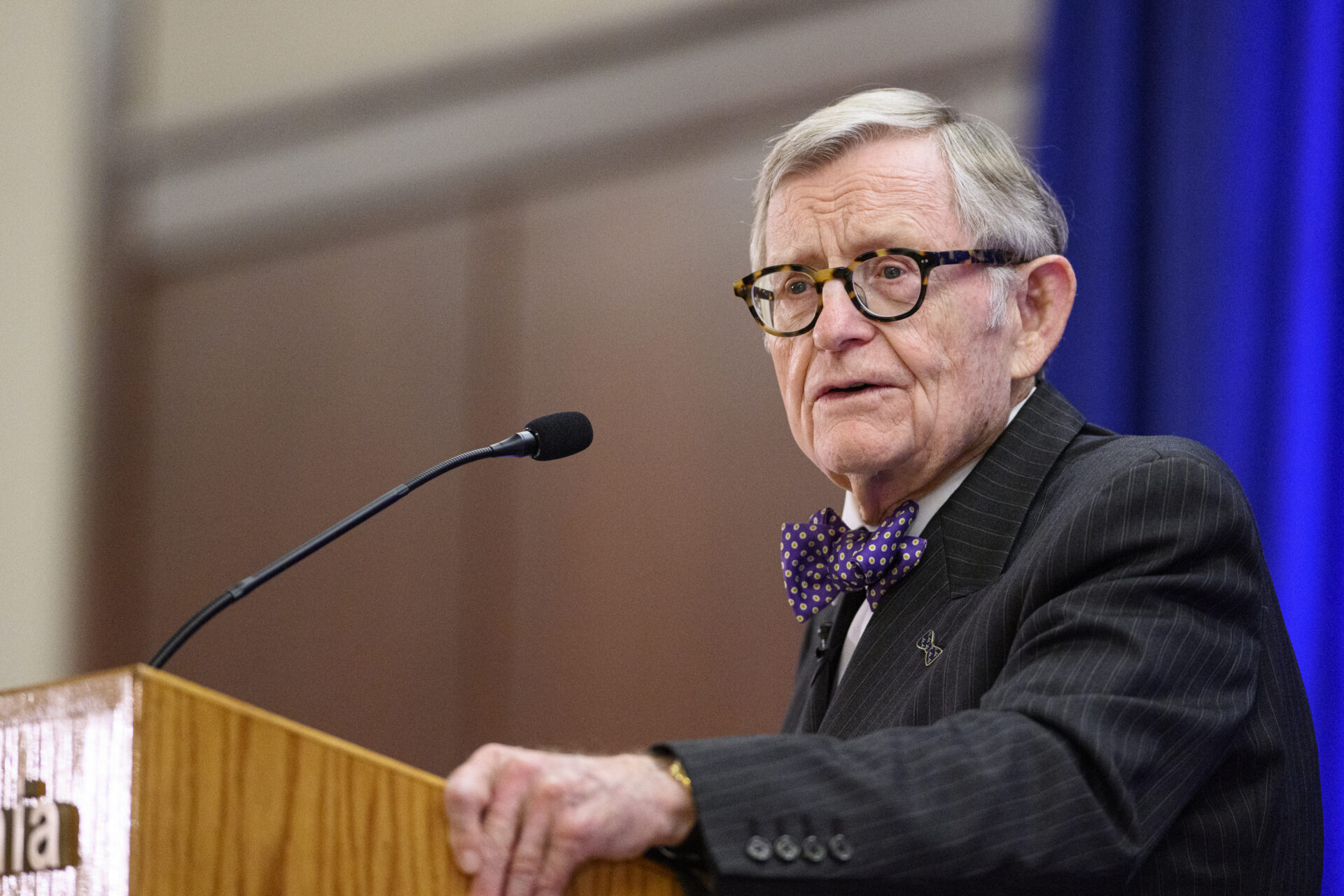 Gordon Gee stands with his hand on the side of a wooden lectern. He wears a black suit with a white shirt, dark purple bowtie and glasses. A microphone just into the center of frame in front of his face