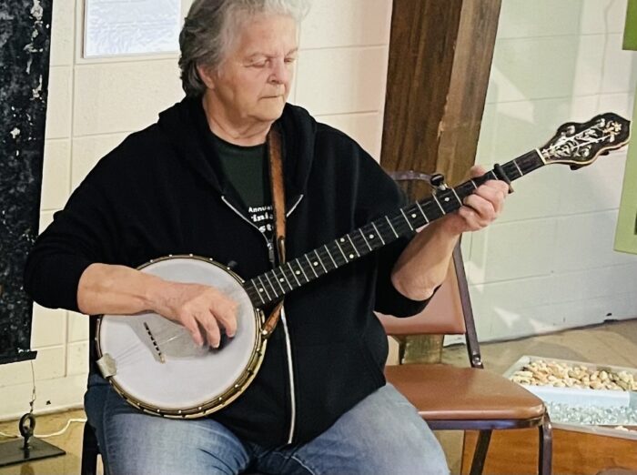 Woman with short grey hair plays the banjo. Her gaze is on the fret board.