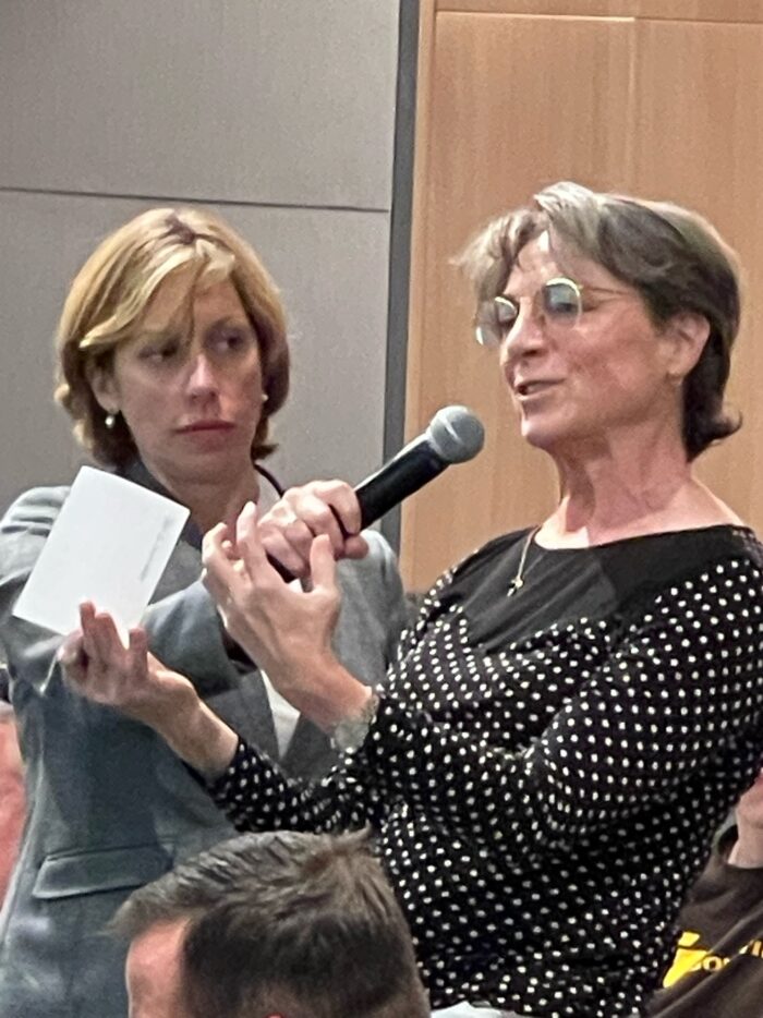 A woman wearing a black and white polka dot shirt and glasses speaks into a microphone that's held by a woman in a business suit.