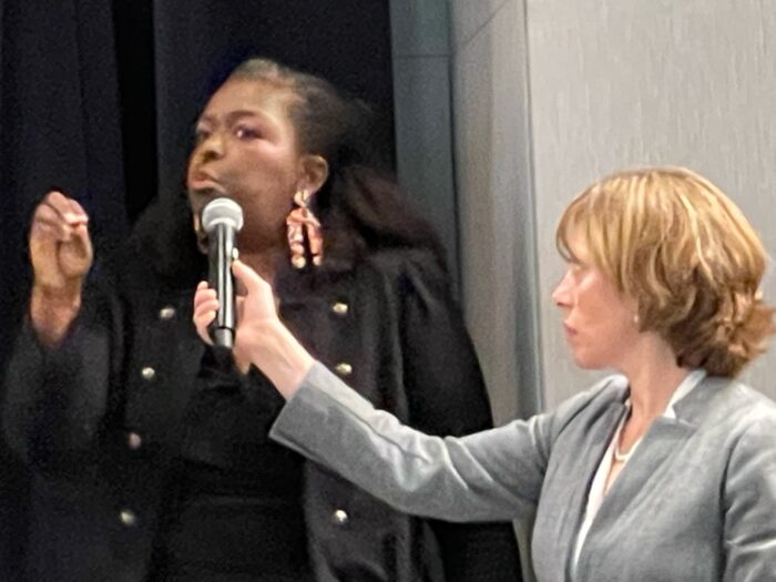 A woman with dark hair and big earrings speaks into a microphone that's held by a woman in a business suit.