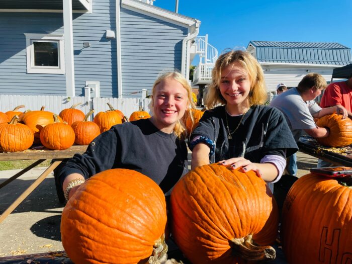 Two girls stand side by side carving pumpkins.