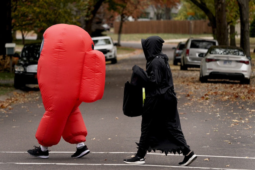 Two trick-or-treaters walk across the street. One kid is dressed in a red blow up suit, while the second kid is dressed up like the grim reaper. There are fall leaves on the ground in the background near parked cars.