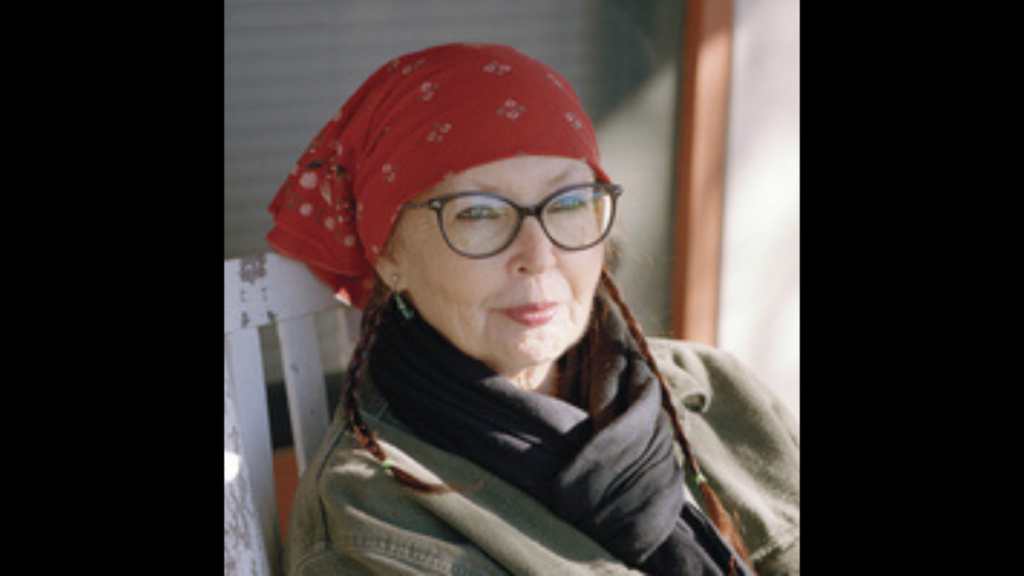 A photograph of an older woman wearing glasses and a red bandana on her head. She has a tan scarf around her neck.