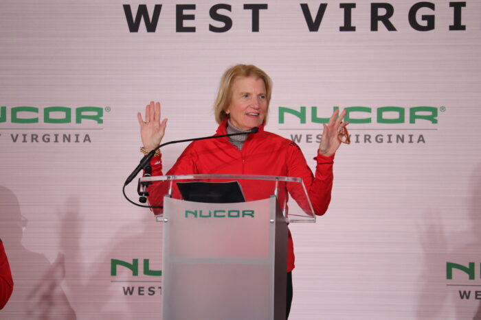 A woman with blonde hair and wearing a red jacket gestures to an audience inside a white tent while holding her glasses.