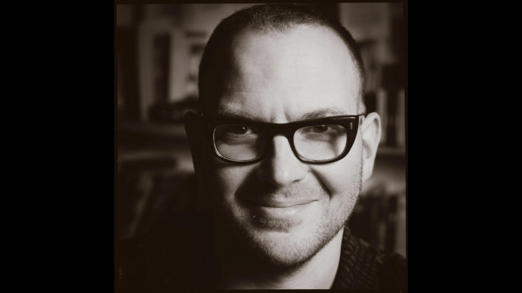 A black and white photo of a headshot of a man wearing black glasses and smiling at the camera.
