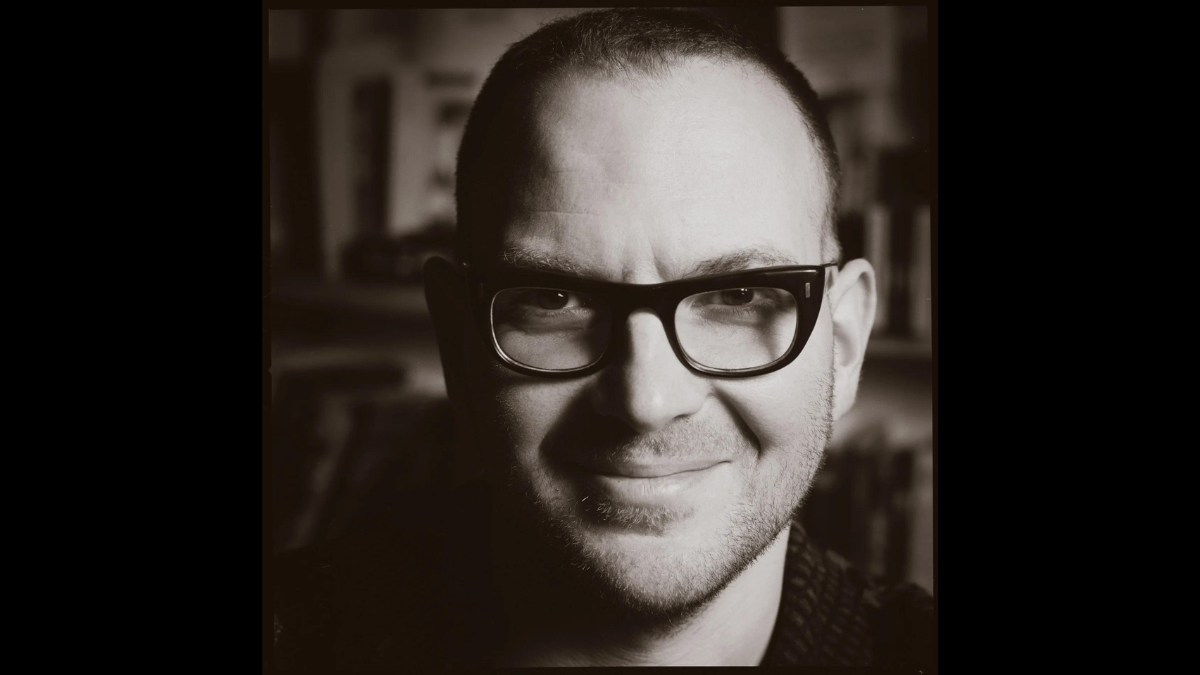 A black and white photo of a headshot of a man wearing black glasses and smiling at the camera.
