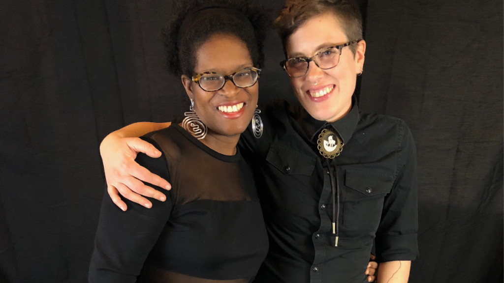 Two people pose for a photo. Both wear glasses and black outfits, smiling for the camera.