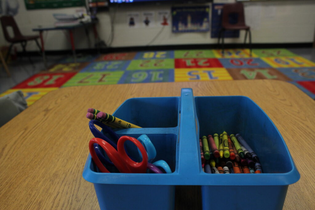 A container of art and school supplies sits on a table in a first grade classroom. In the background a colorful carpet with numbers on it can be seen.