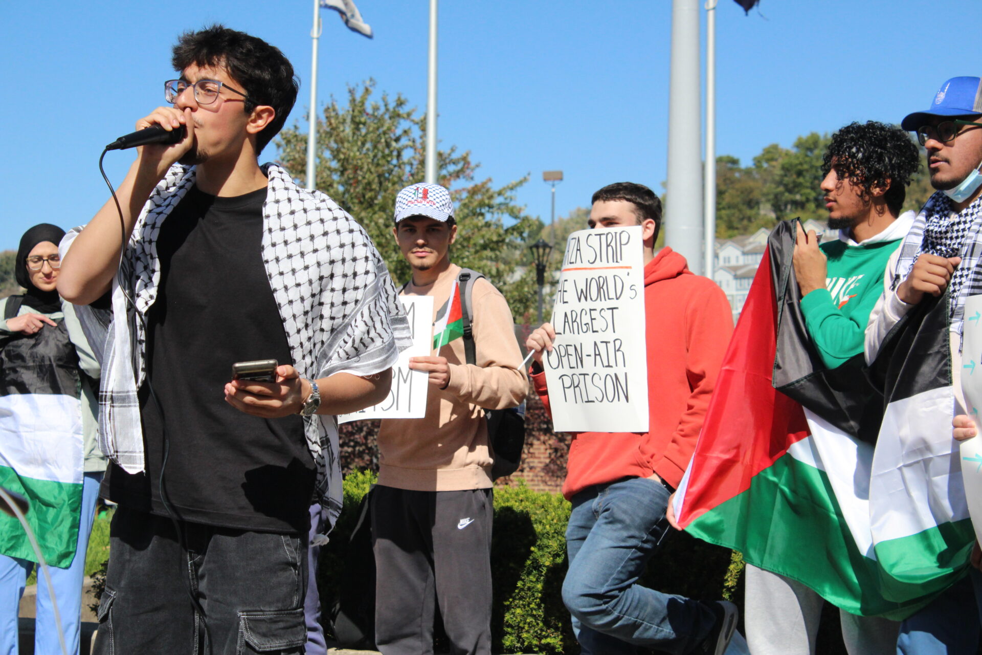 A man wearing a black shirt, black jeans and with a black and white scarf - known as a Palestinian keffiyeh - over his shoulders, speaks into a microphone while holding a cell phone. Behind him stand men holding a Palestinian flag, while a man in red holds a sign that reads "Gaza Strip: The World's Largest Open-Air Prison"