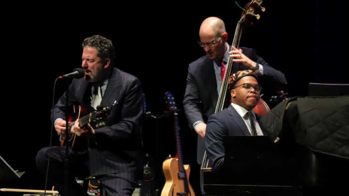 A man sings and plays the guitar next to a man in a hat playing the piano and a man playing the upright bass.