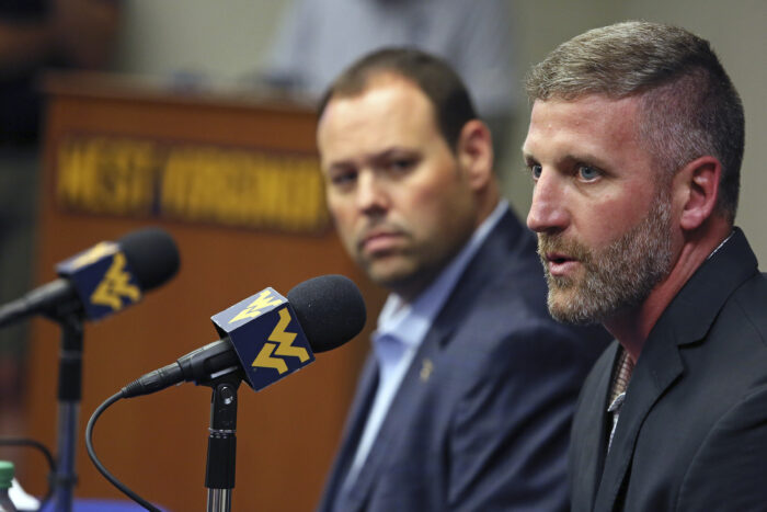 Two men sit at a table and speak into microphones for a press conference.