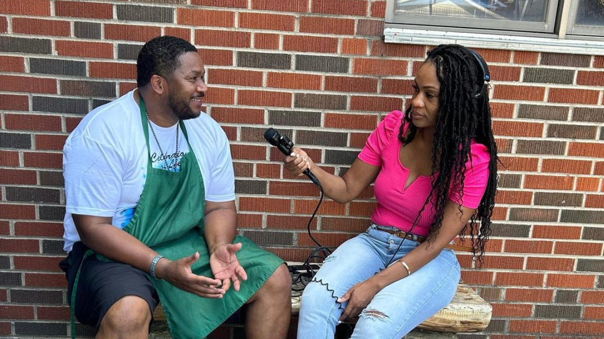 Two people sit on a bench. A man and a woman. The man wears a green apron. The woman wears a pink shirt. She holds a microphone up to the man.