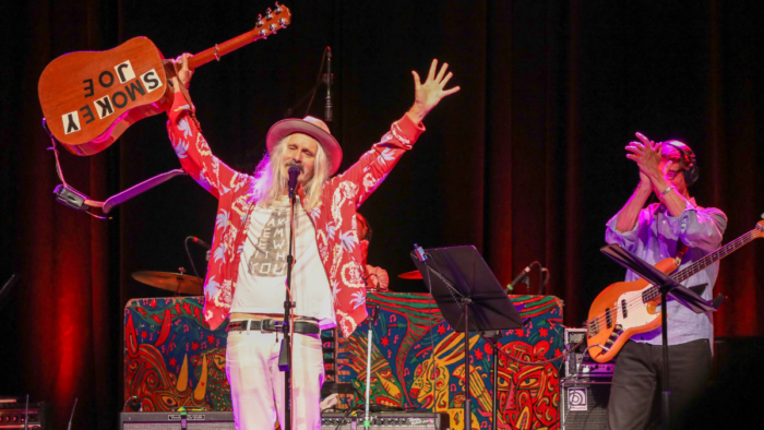 A man with a hat, long grey hair, a red jacket and white pants holds his guitar above his head in his right hand and his left arm is raised above his head.