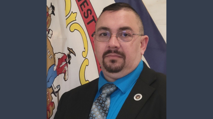 A man with a short haircut, wearing glasses and a black suit with a blue shirt and grey tie.