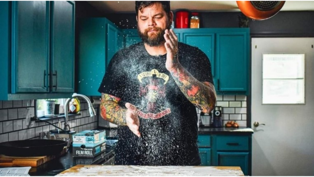 Bearded man in kitchen wearing black logoed t-shirt dusting flour in the air