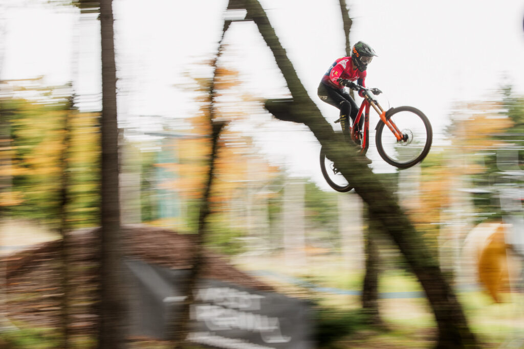 A mountain biker flying through the air is the only thing in focus as they come off a jump between trees. The rider wears a red top, black pants and a black helmet while riding an orange bike. Below their rear tire a "West Virginia" sign is blurred by the photo's motion.