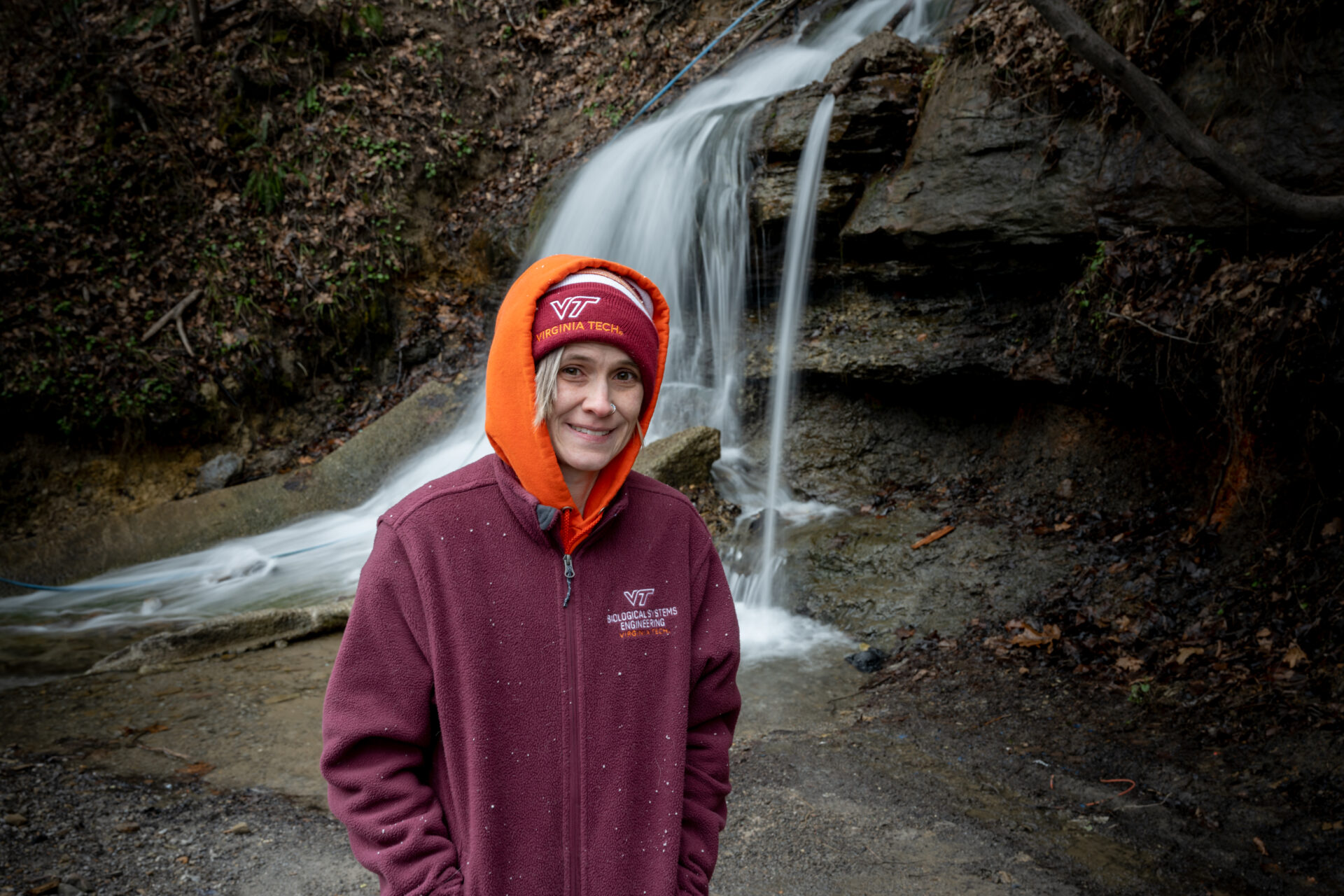 A woman in a maroon hoodie standing in front of a small waterfall smiling toward the camera.