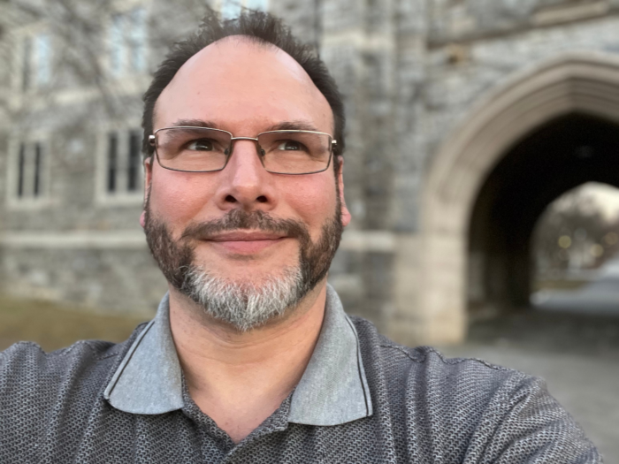A man with dark, thinning hair and in glasses looks off camera. He wears a gray polo shirt.