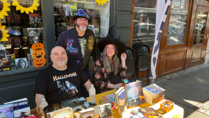 Three people sit behind a table stacked with horror and sci-fi books. They smile for the camera and are dressed up for Halloween.