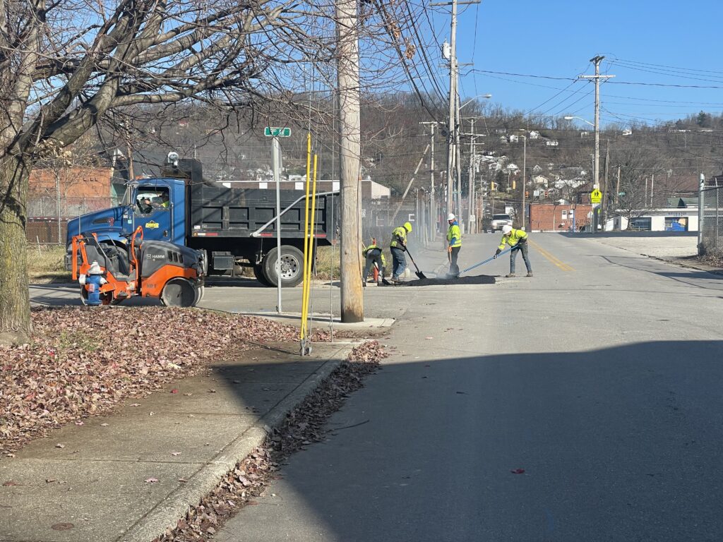 Crews work shoveling black gravel along a roadside.