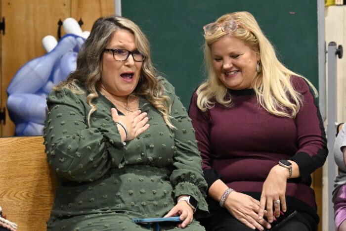 A woman wearing a green dress clutches her chest in surprise as a woman wearing a maroon top looks on.