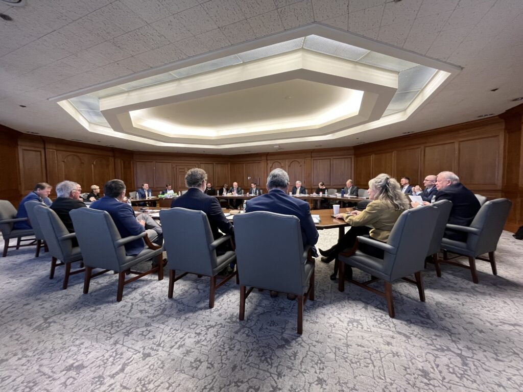 A group of people dressed in suits sit at a board meeting table.