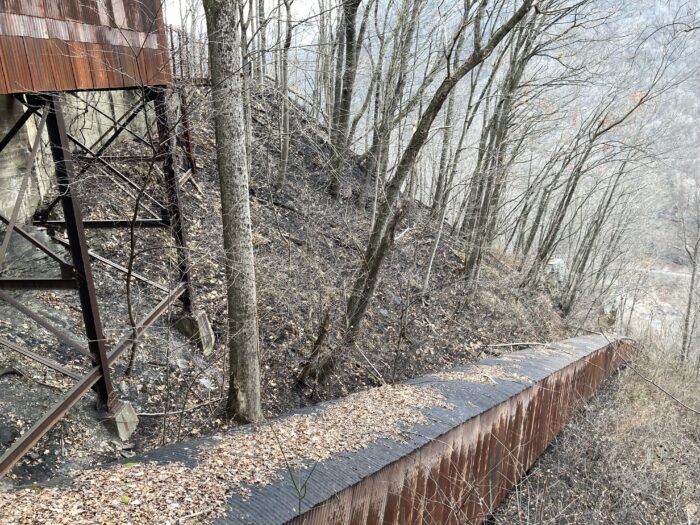 A rusty looking wall lines a path where the trees and earth is burned.