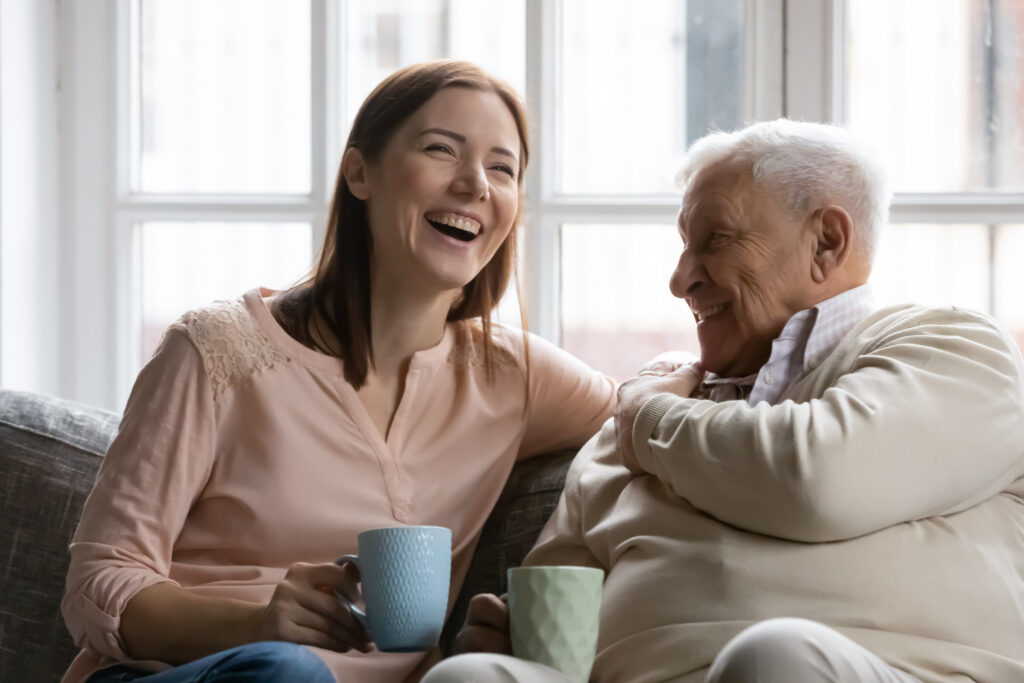 An older father sits next to his grown daughter on a couch. Both hold a coffee mug and are laughing.