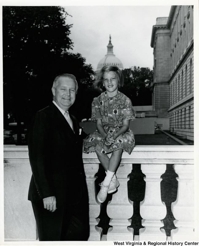 A black and white photo featuring an older man in a black suit standing next to a little girl who sits on white railing. Both smile for the camera. The U.S. Capitol building can be seen in the distance.