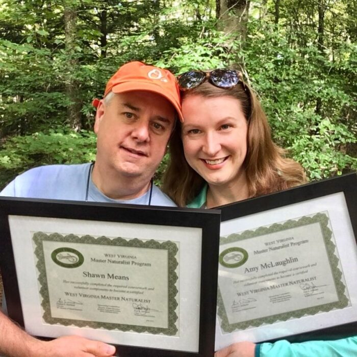 Two middle-aged people, a man and a woman, stand in front of trees holding framed certificates that read "West Virginia Master Naturalist Program."