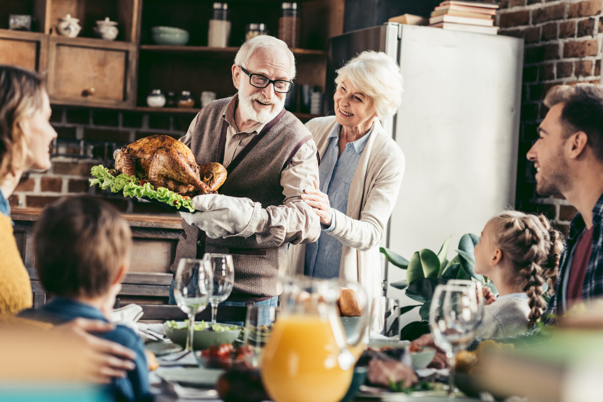 A grandfather and grandmother serve a turkey at the dinner table to their family. The elderly couple are smiling.