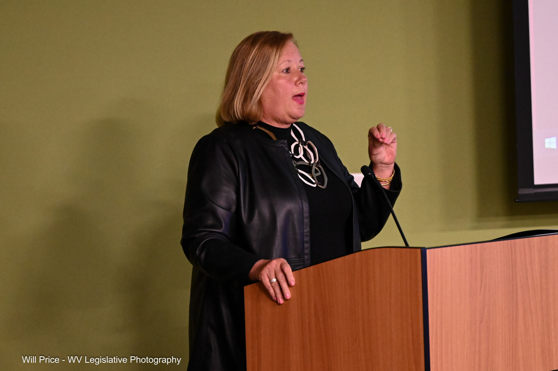 A woman wearing black business attire speaks from a podium in front of a green wall.