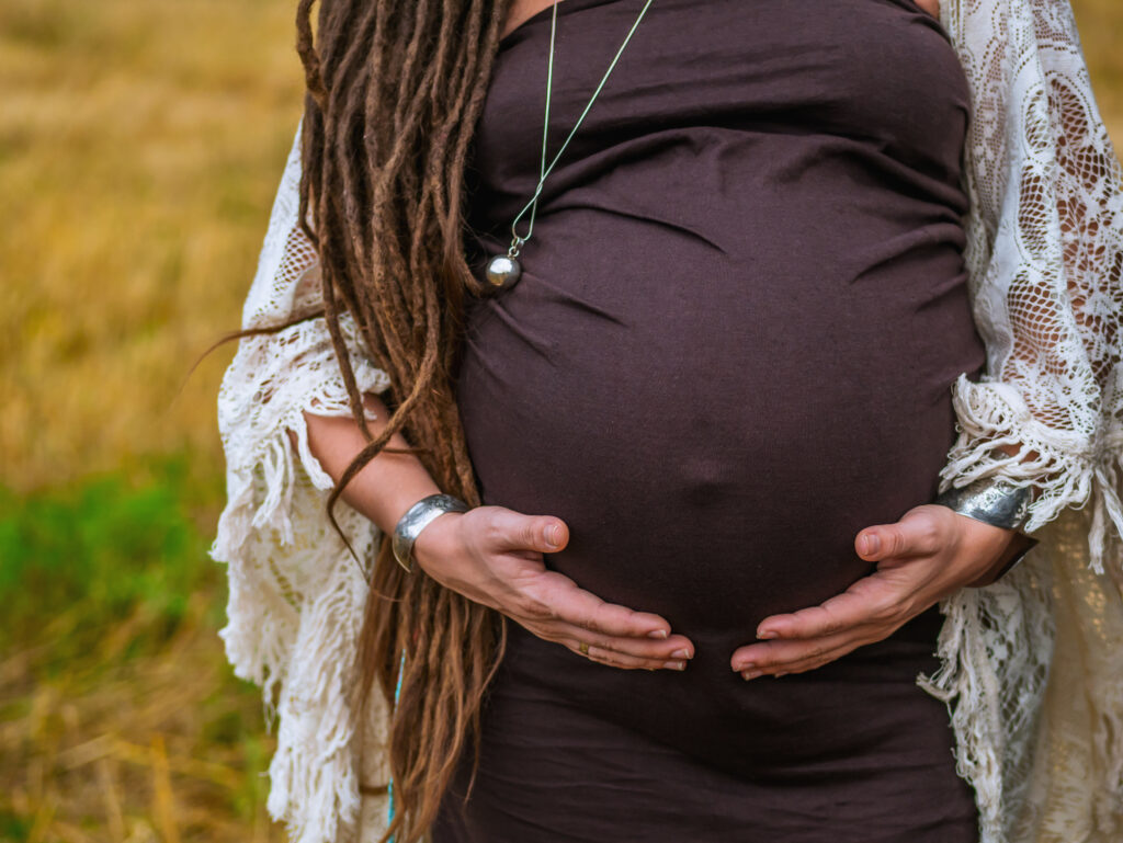 A pregnant woman stands in a field, cradling her baby bump. She is wearing brown and white.