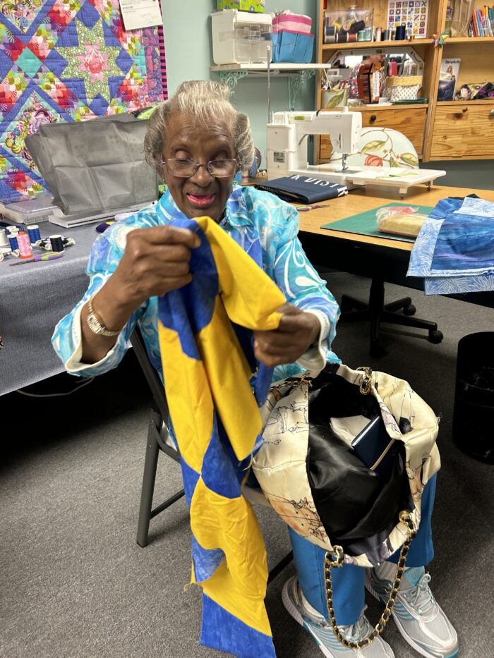 An elderly woman with glasses sits in a chair and holds up blue and yellow fabric.