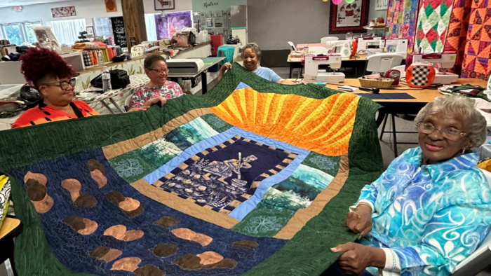 Four women hold up a large quilt, smiling.